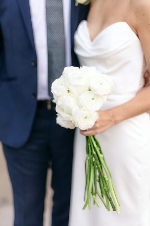 Elegant white rose bridal bouquet with long stems held against a white wedding dress next to a navy suit.