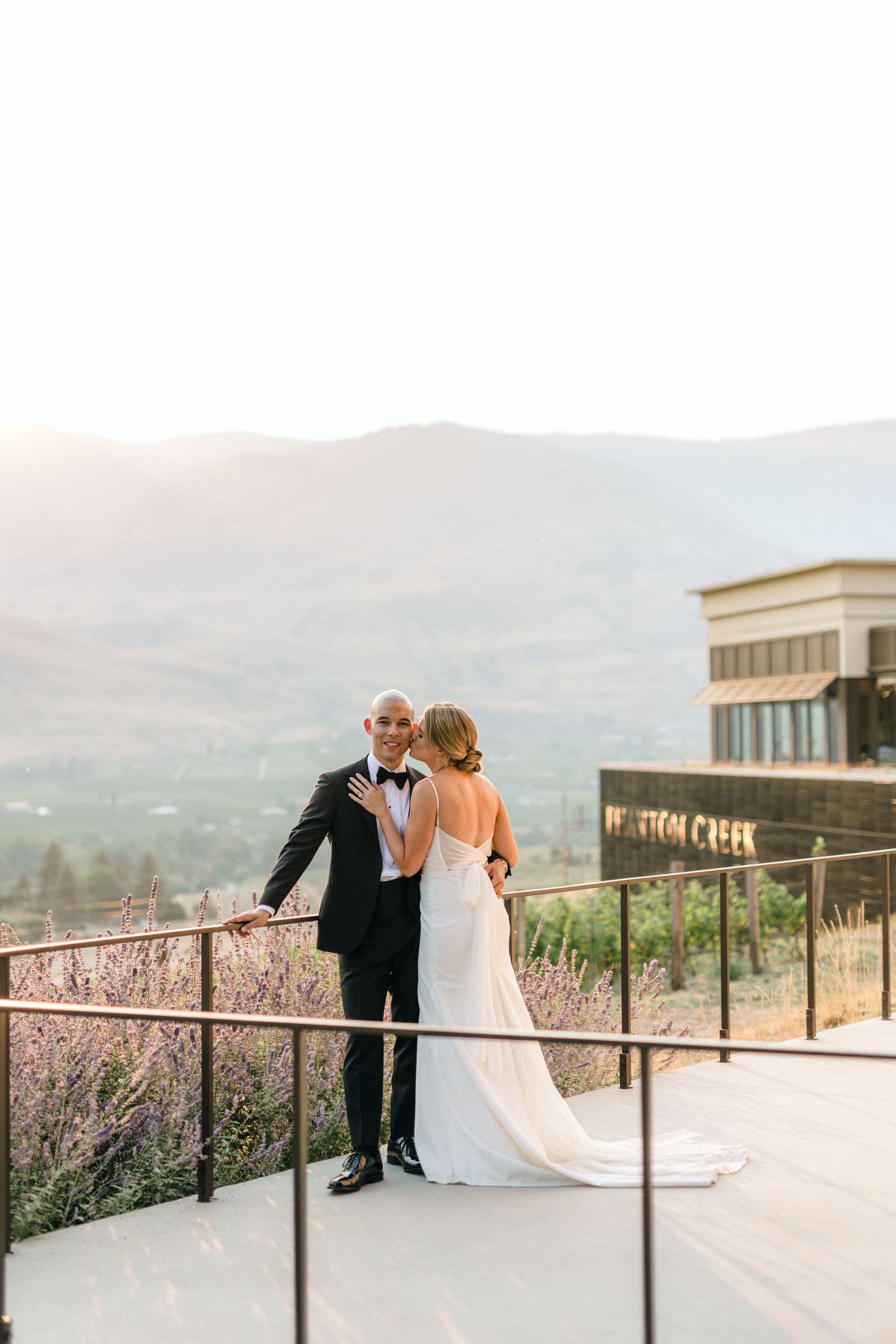 Wedding couple posing along railing with scenic mountain views.