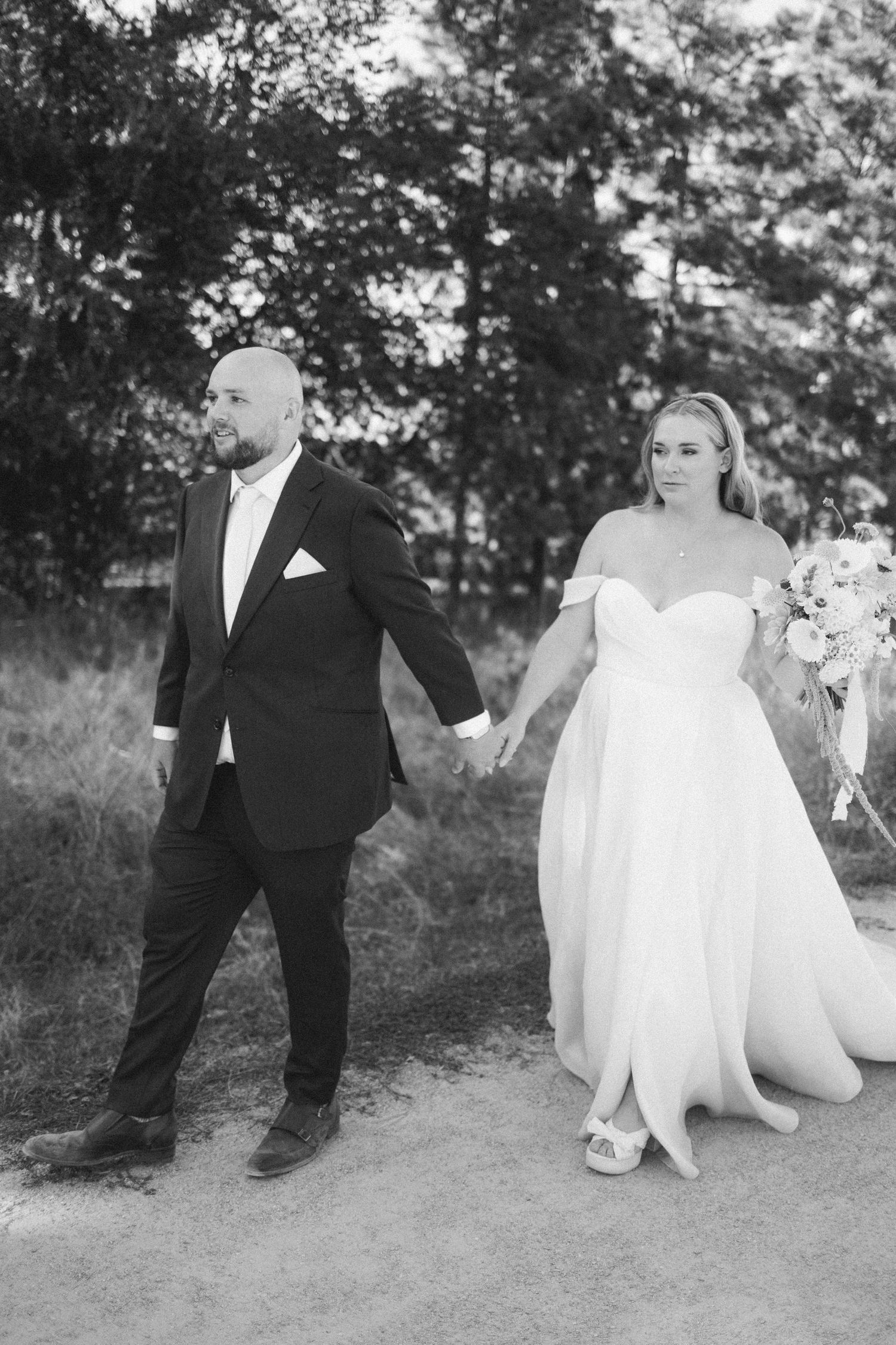 A black and white photo of a bride and groom holding hands while walking outdoors, with trees in the background.