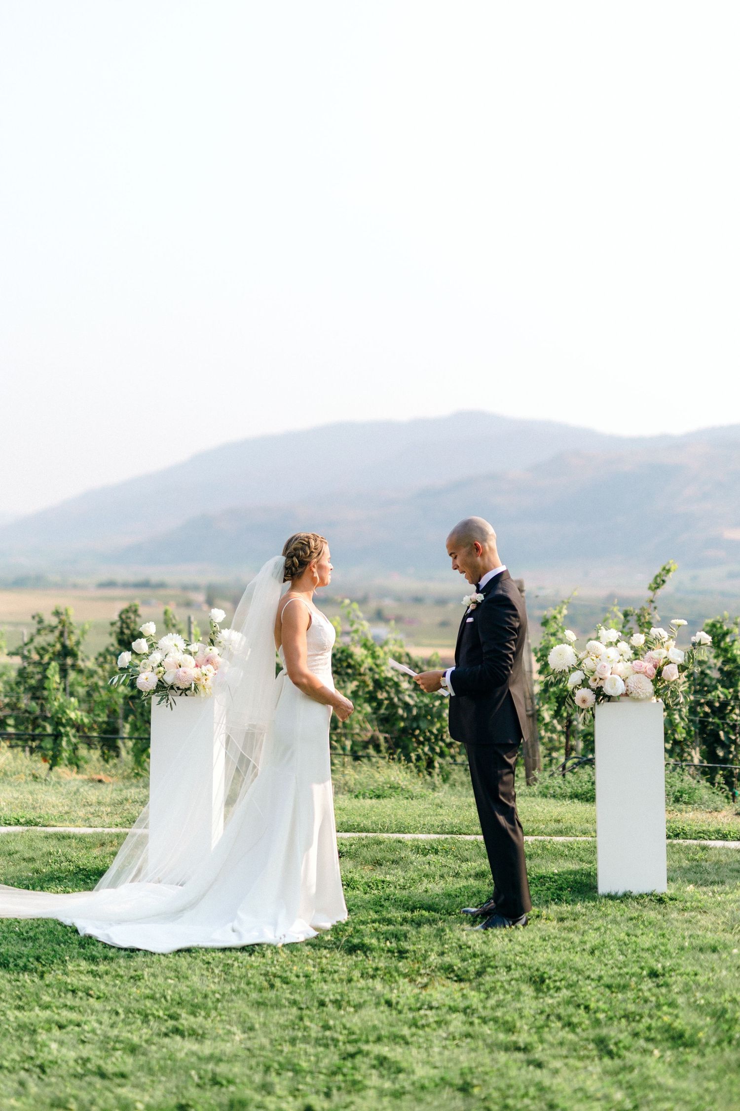 Wedding ceremony setup in a vineyard with white floral arrangements and mountain backdrop.
