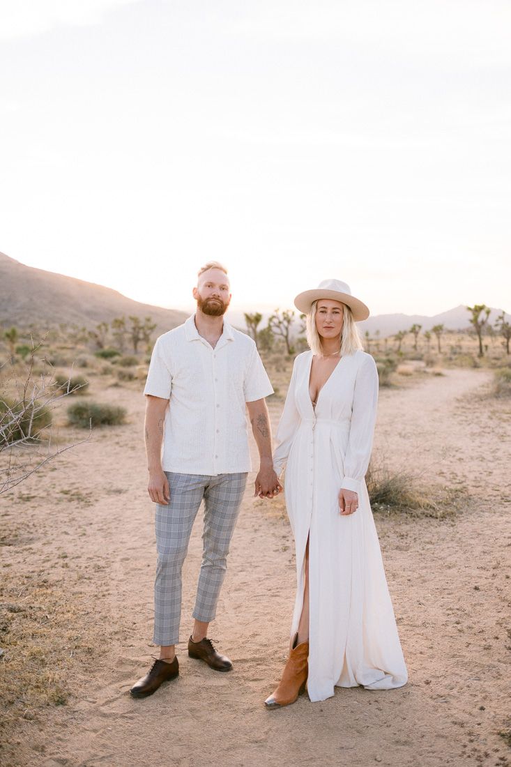 Fashion-forward couple in white outfits pose together in a desert setting during golden hour.