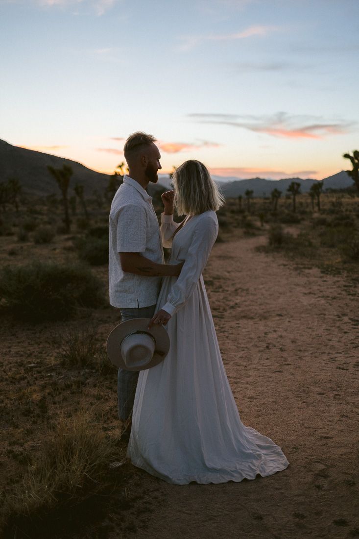 A couple shares a romantic moment in the desert at sunset with mountains in the background.