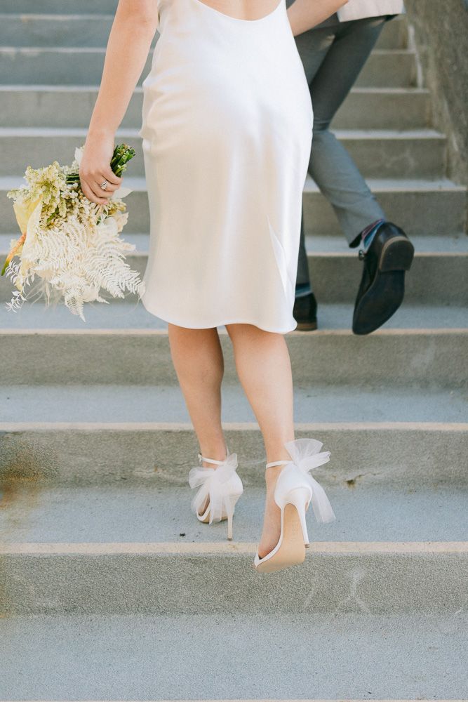 White wedding dress and heels with bow details ascending concrete stairs holding a delicate bouquet.