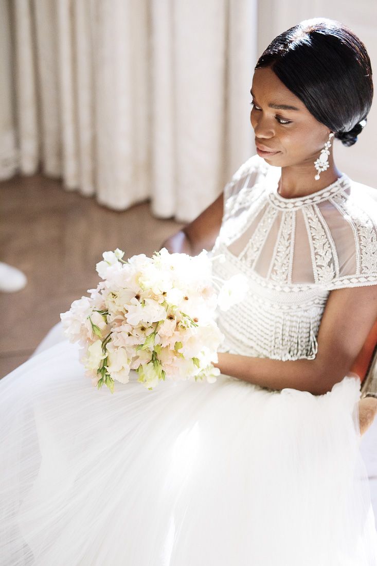 Bride in detailed lace bodice gown holds pastel bouquet while seated.