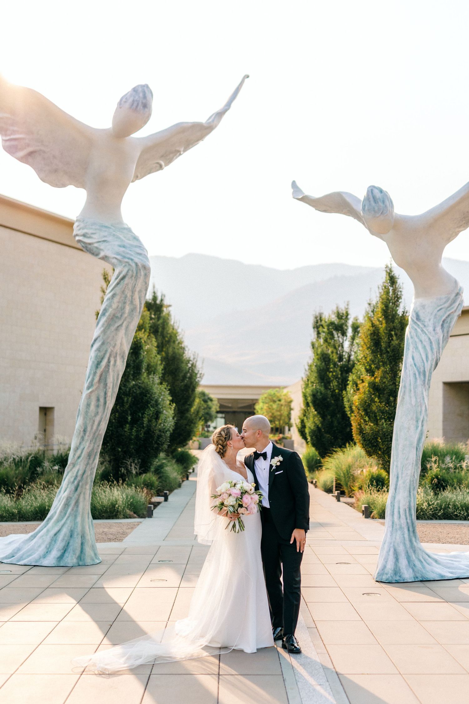 A couple poses for a wedding photo between two large angel wing sculptures at an elegant outdoor venue.