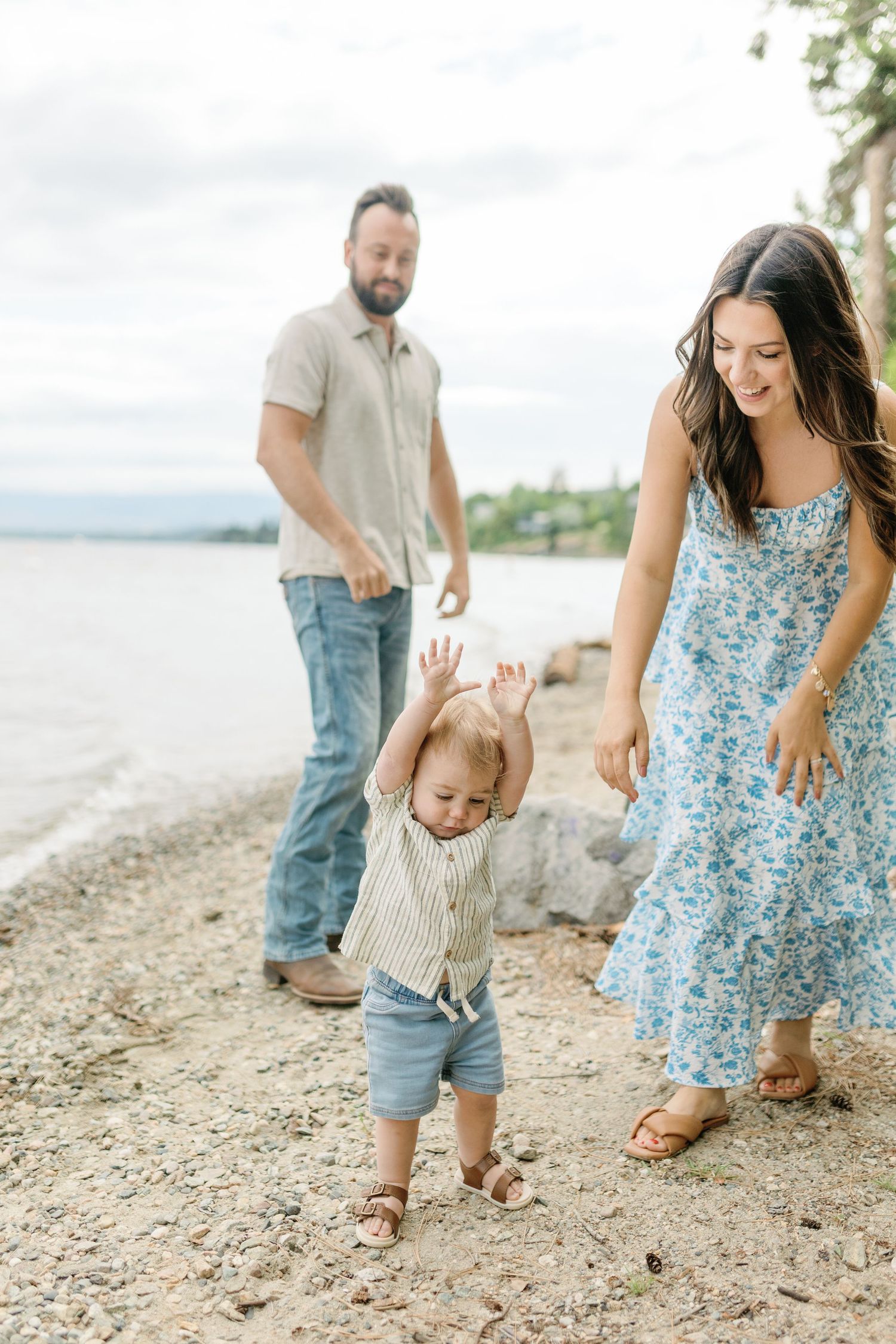 A family walks together on a rocky beach shore, with a child in a blue floral dress and casual summer attire.