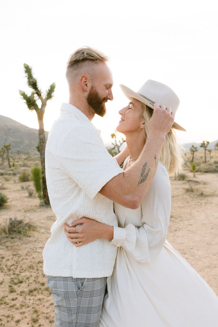 Couple in white clothing embrace and laugh during desert photoshoot with Joshua tree in background.