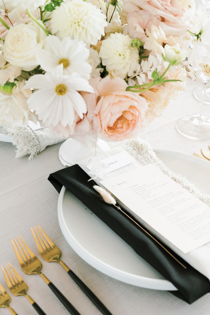 Place setting with white plates, gold flatware and blush floral arrangements on white tablecloth.