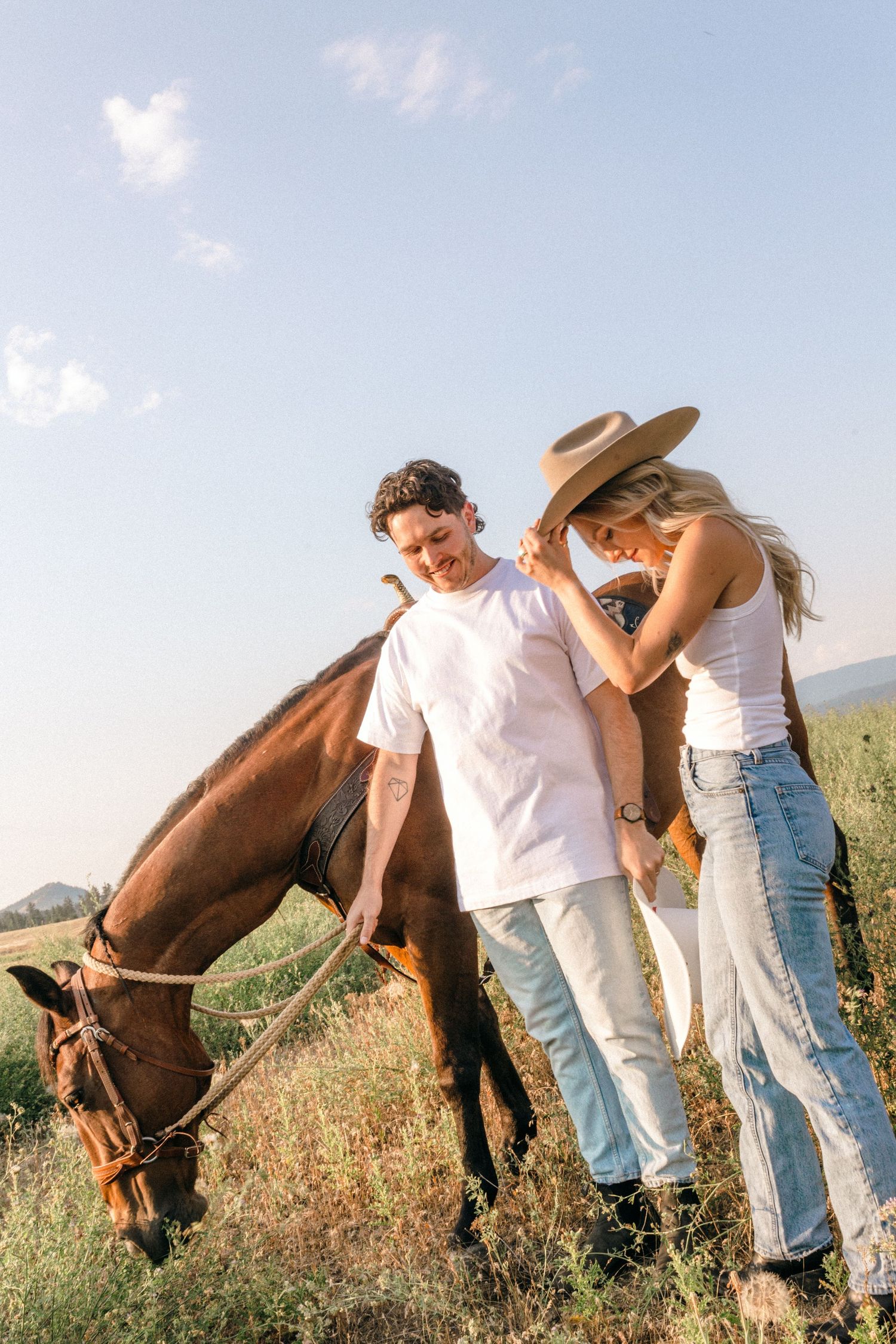 Couple in Western wear spending time with their brown horse in a sunny mountain meadow.