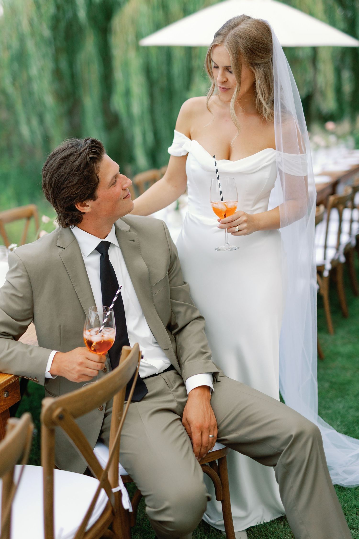 A bride and groom share a romantic moment in an outdoor garden setting with white umbrellas and beige suits.