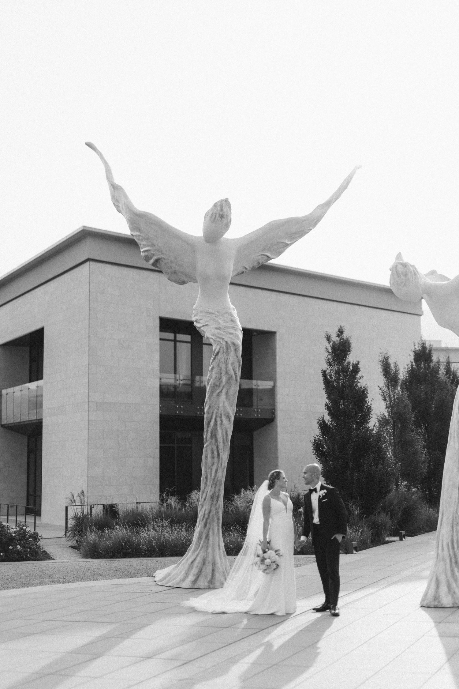Black and white wedding photo with tall angel statues framing the couple at an elegant venue.