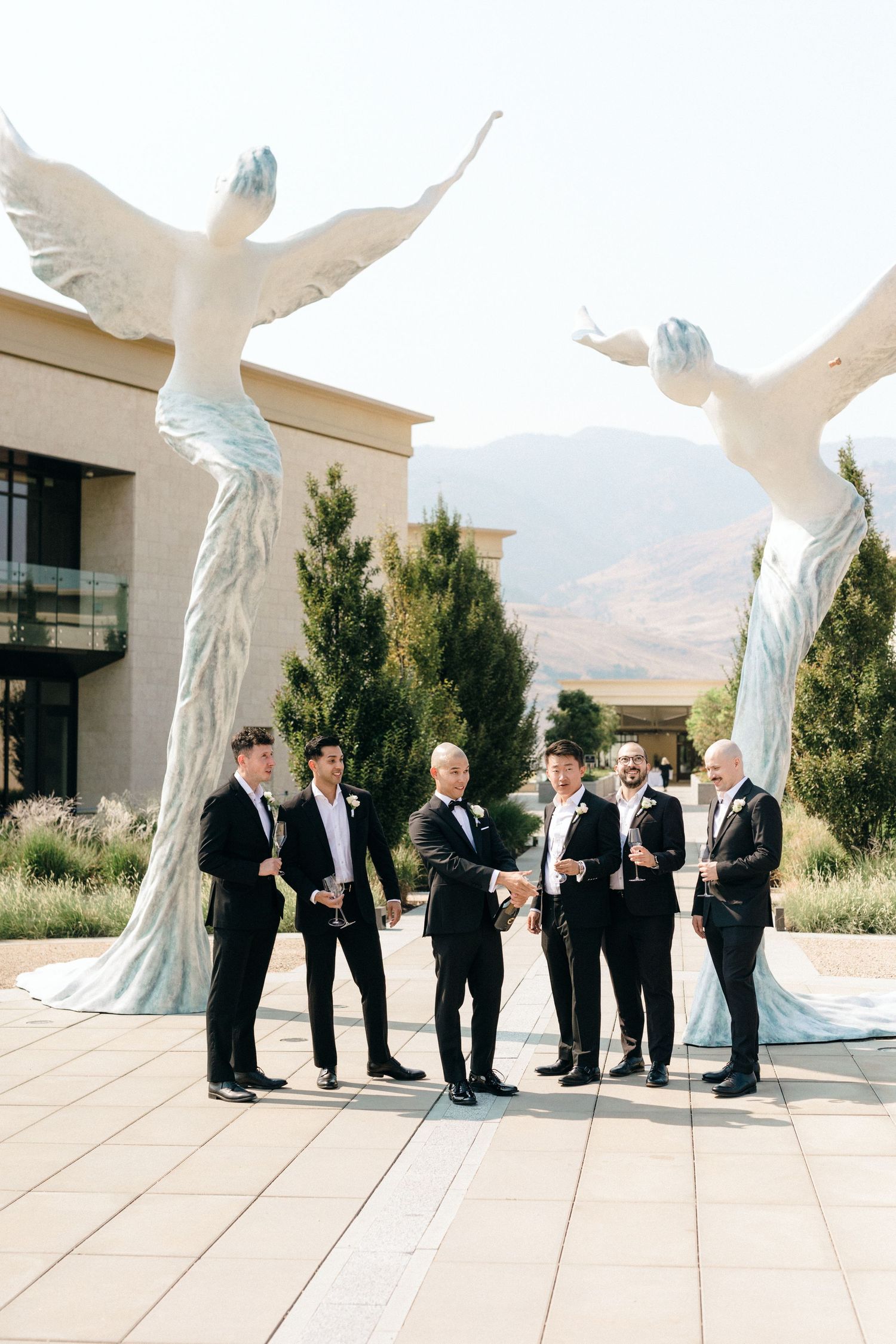 Groomsmen in black suits gather for a photo between two towering white angel sculptures at an outdoor venue.