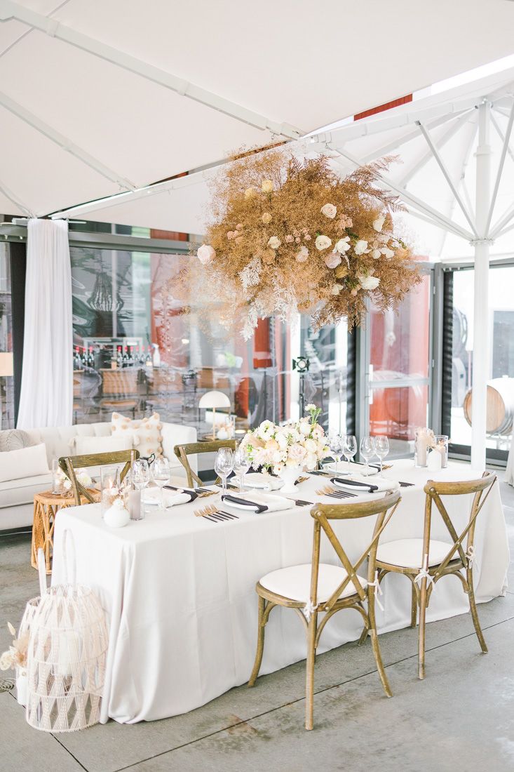 White tablescaped dining setup with dried floral arrangements and gold chairs under a tent structure.