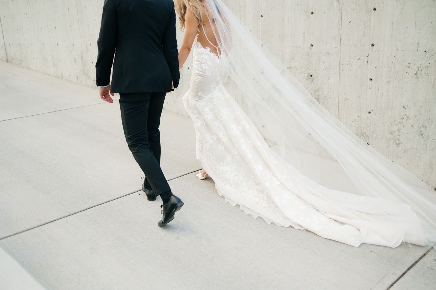 Detail shot of flowing wedding dress train and groom's shoes on concrete floor.