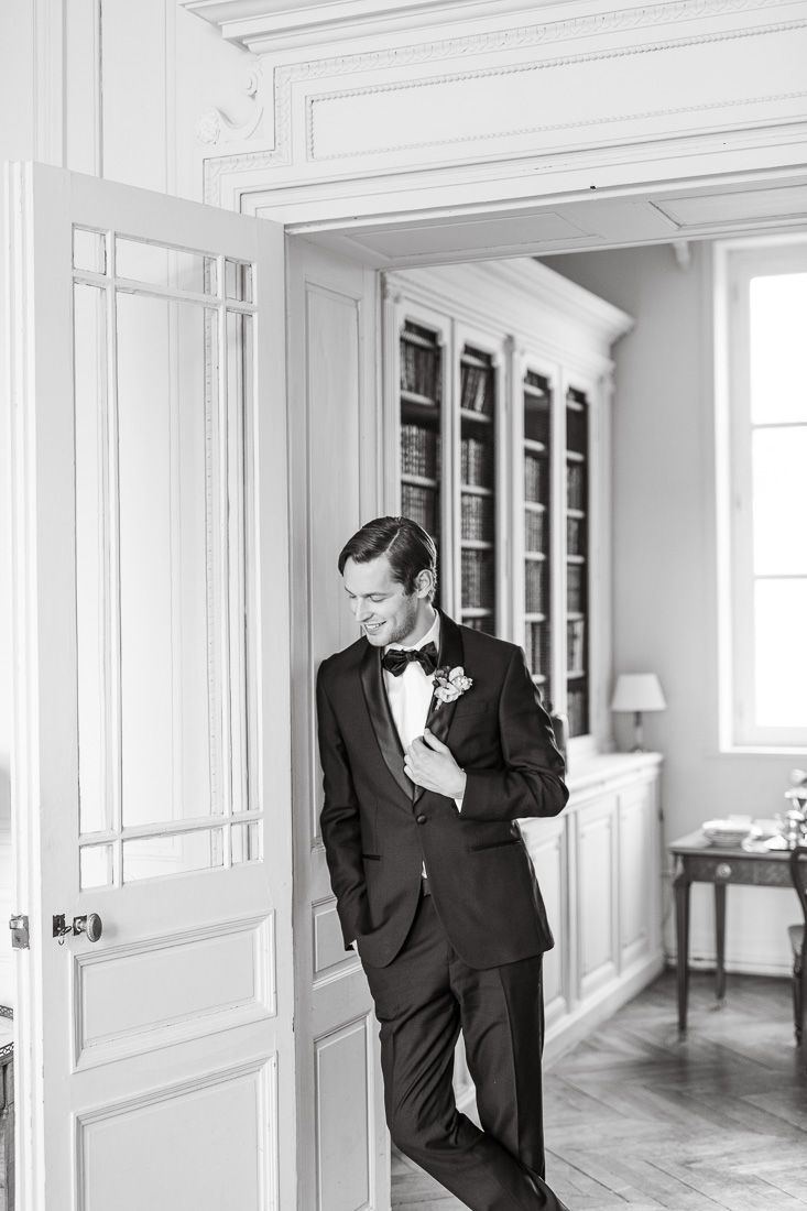 Classic black and white wedding photo showing a groom in a tuxedo leaning against an elegant doorway.