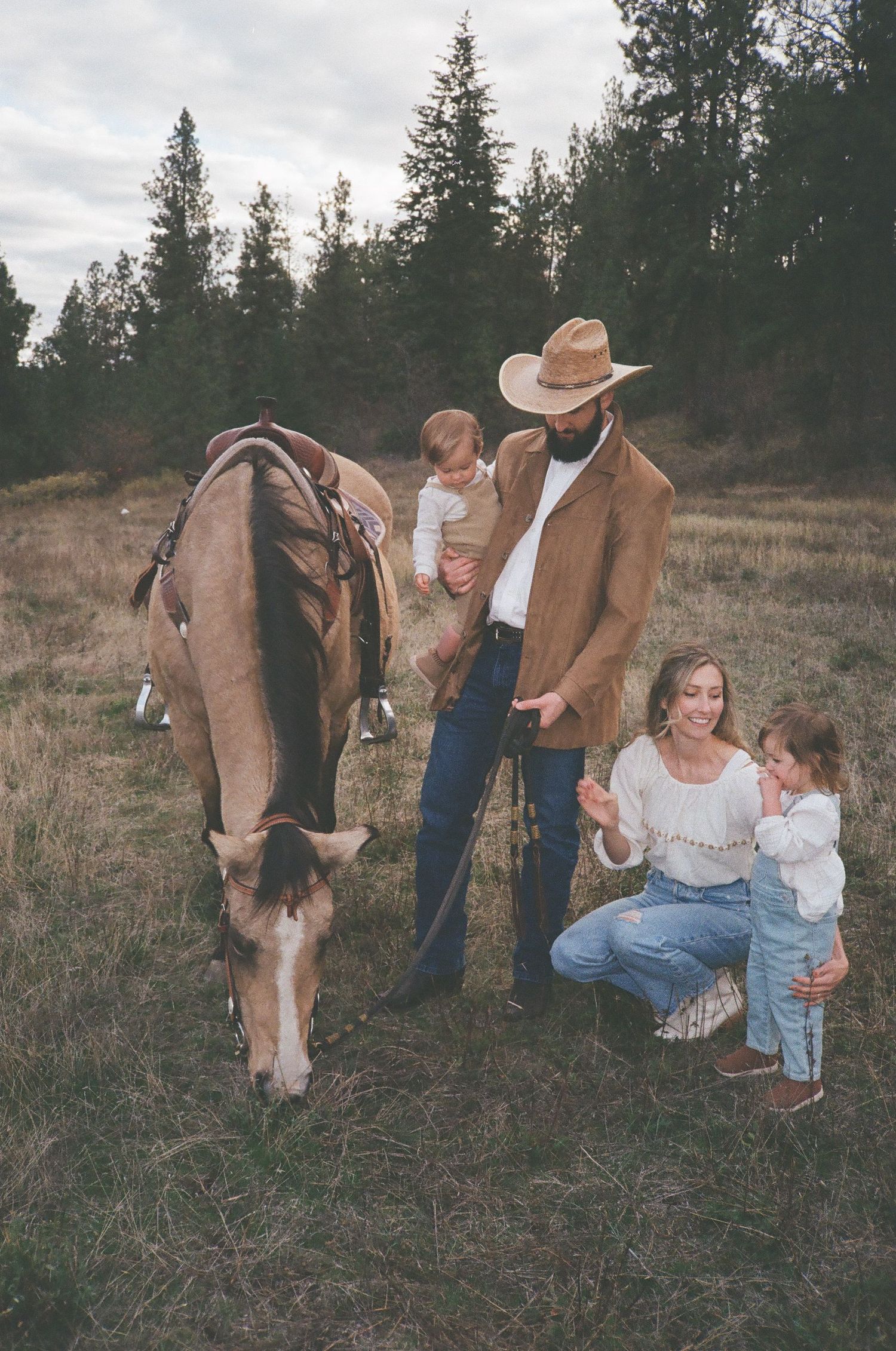 A family in western attire poses with their horse in a field with evergreen trees in the background during autumn.