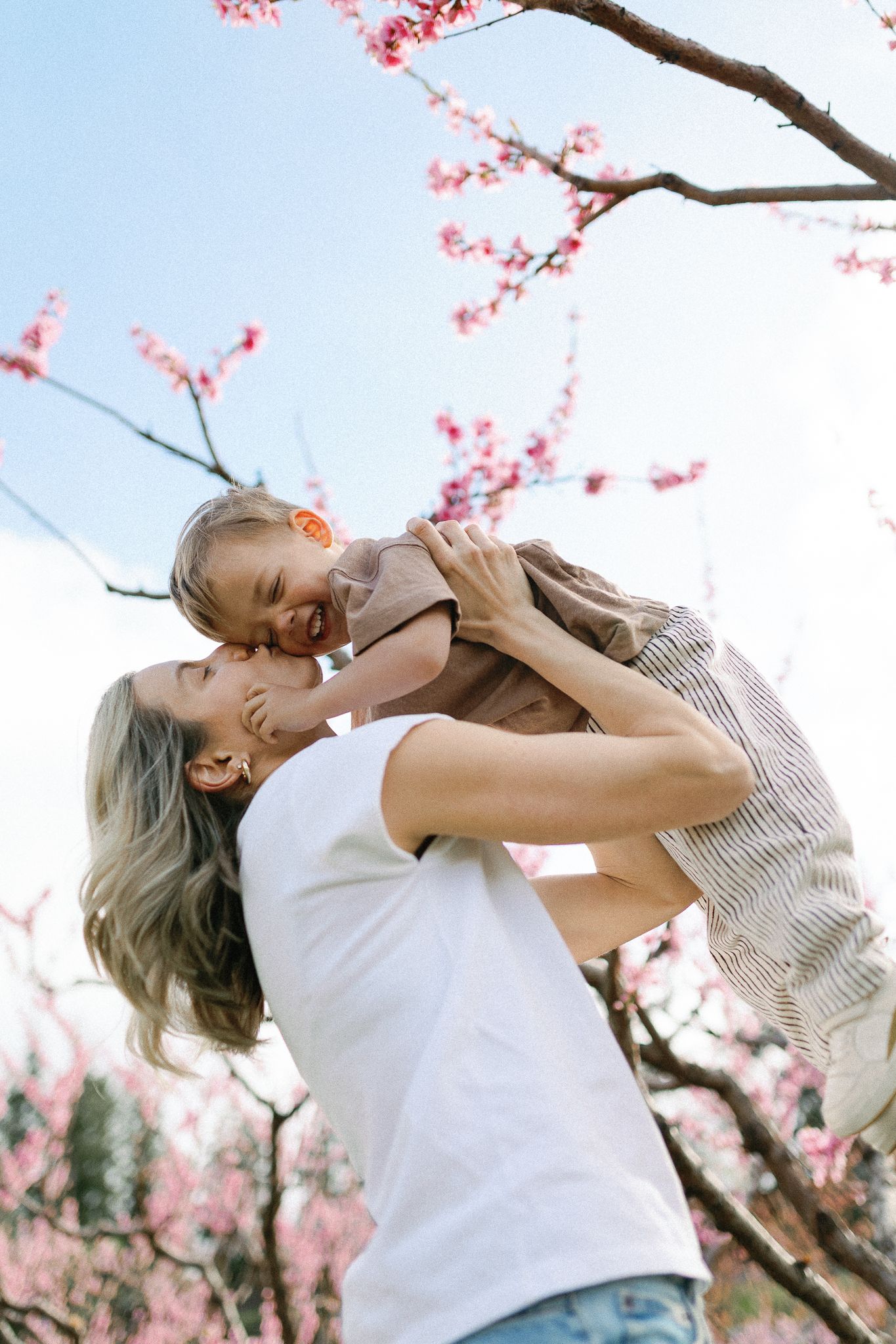 A tender moment under blooming pink cherry blossoms as a caregiver lifts up a child against a spring sky.