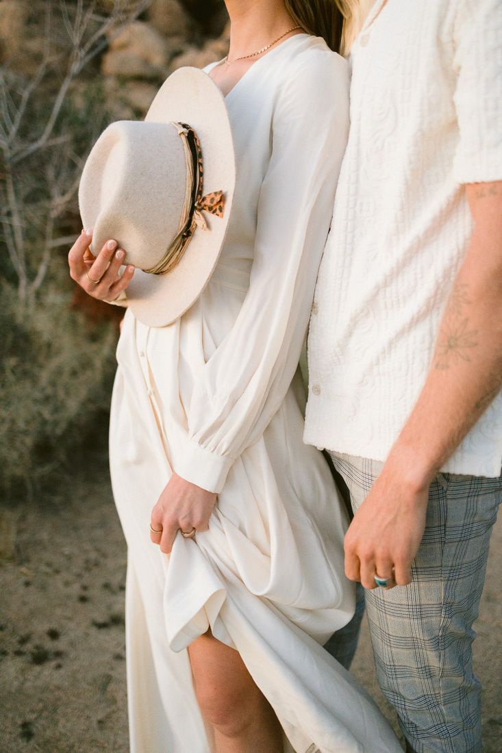 Close-up shot of a couple in white outfits holding a beige hat against a rocky desert backdrop.