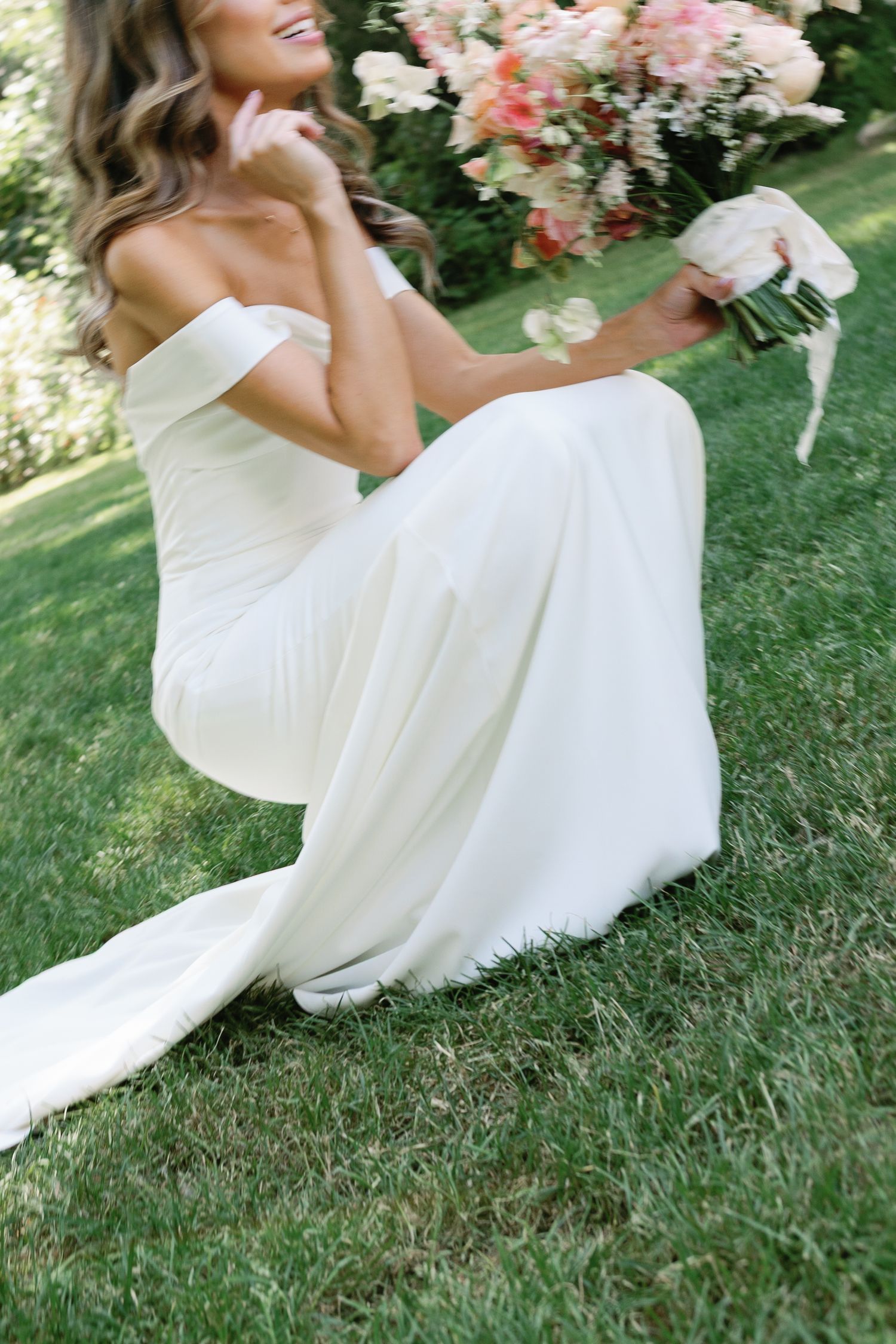 An elegant off-shoulder wedding dress cascades gracefully on lush grass beside pink floral arrangements.