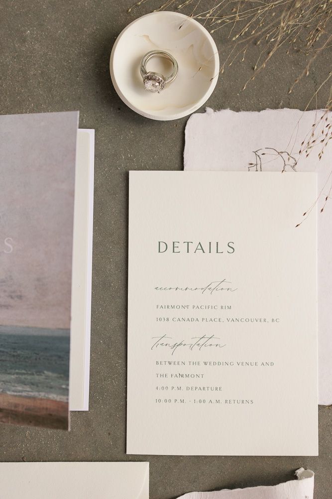 Wedding details card and engagement ring on small dish with coastal artwork and dried flowers.