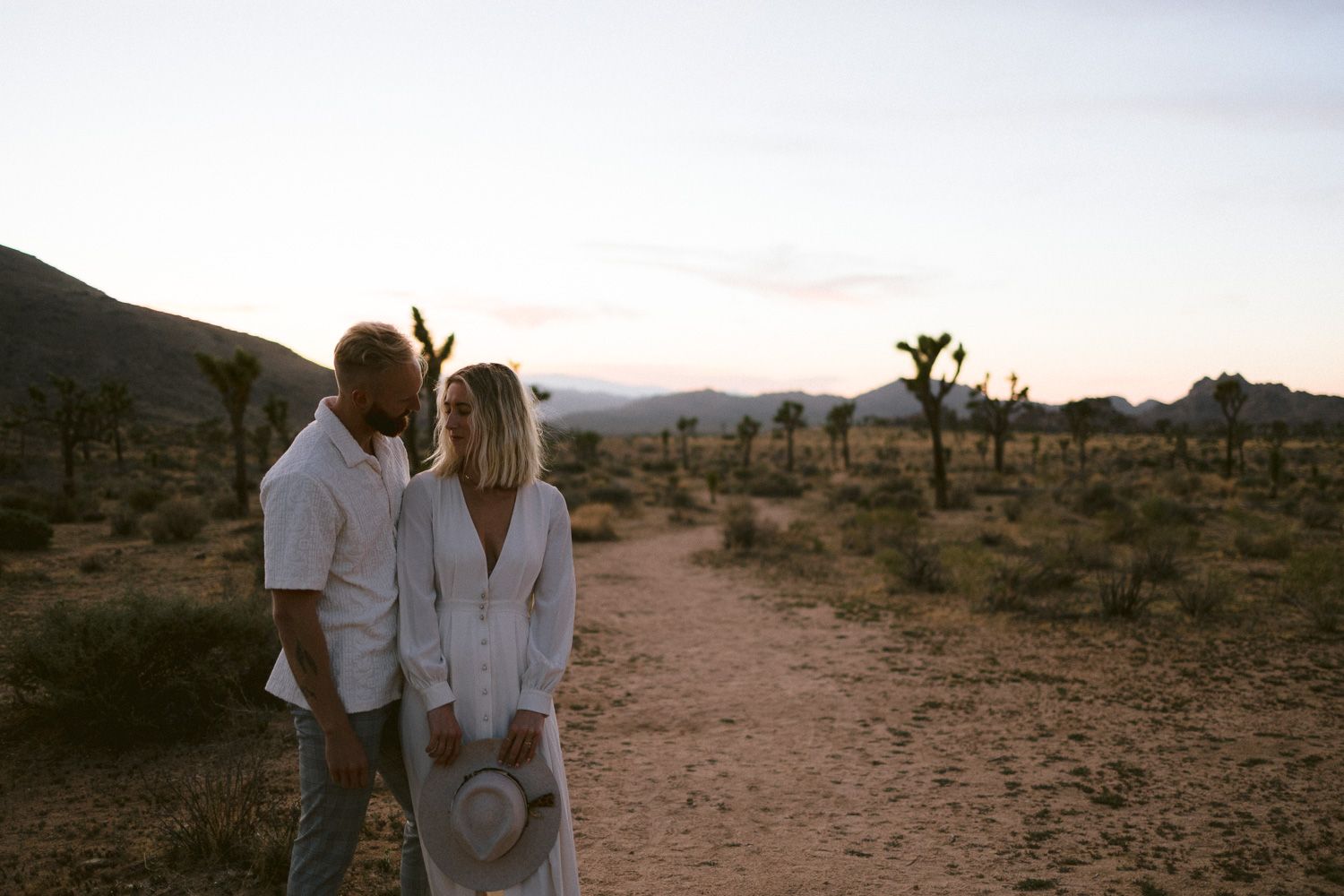 A couple in white clothing embraces on a desert path with Joshua trees at sunset.