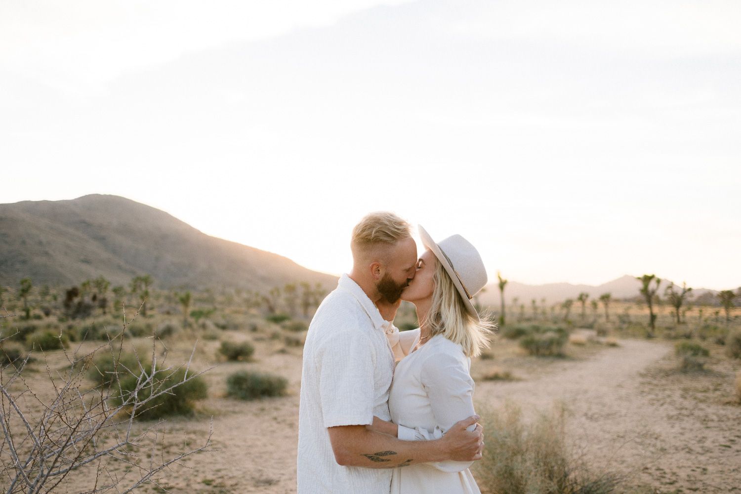 An intimate desert portrait of a couple embracing with Joshua trees and mountains behind them.