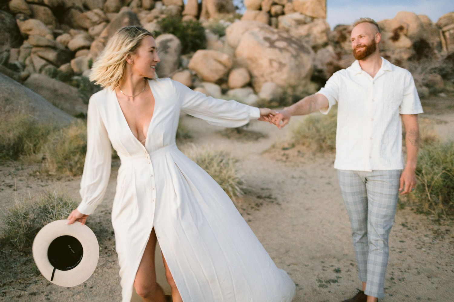 Couple in white outfits holding hands while walking along a desert trail with rocky formations.