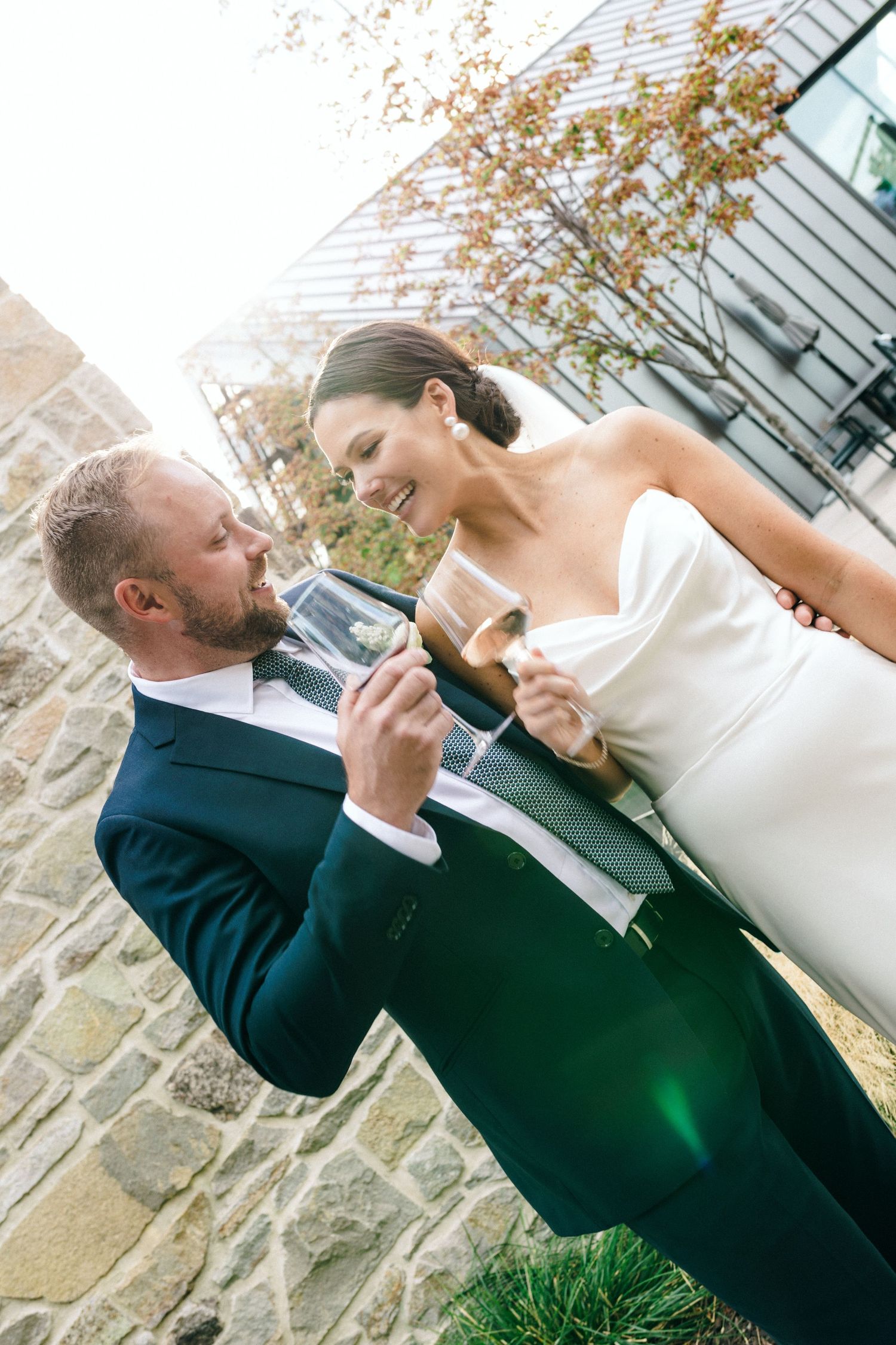 Newlywed couple in formal attire share a playful moment while toasting with wine glasses outdoors.