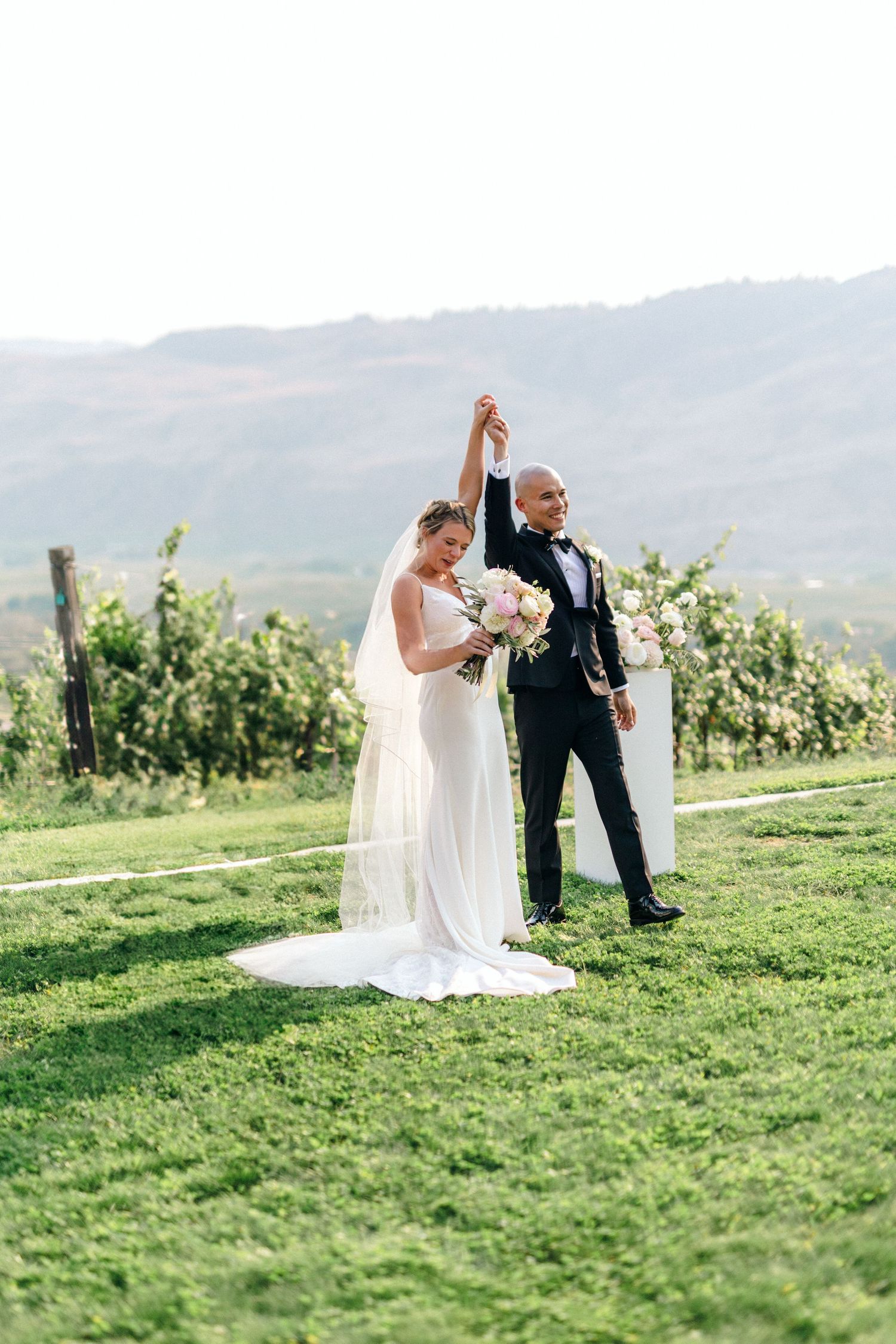 A wedding couple poses together on a grassy hillside with mountains in the background.