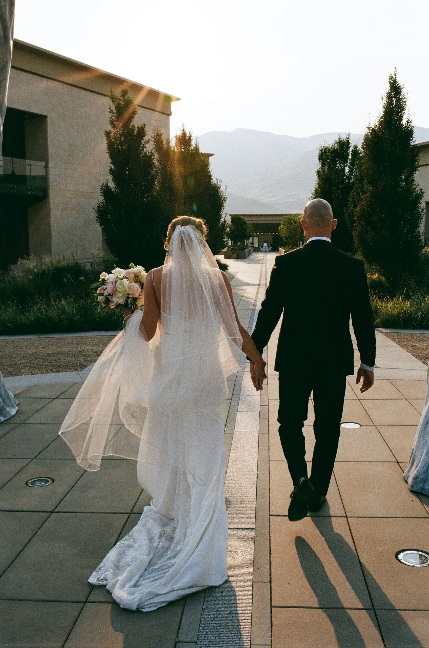 Bride in flowing veil and groom walking hand in hand on outdoor path with mountains in background.