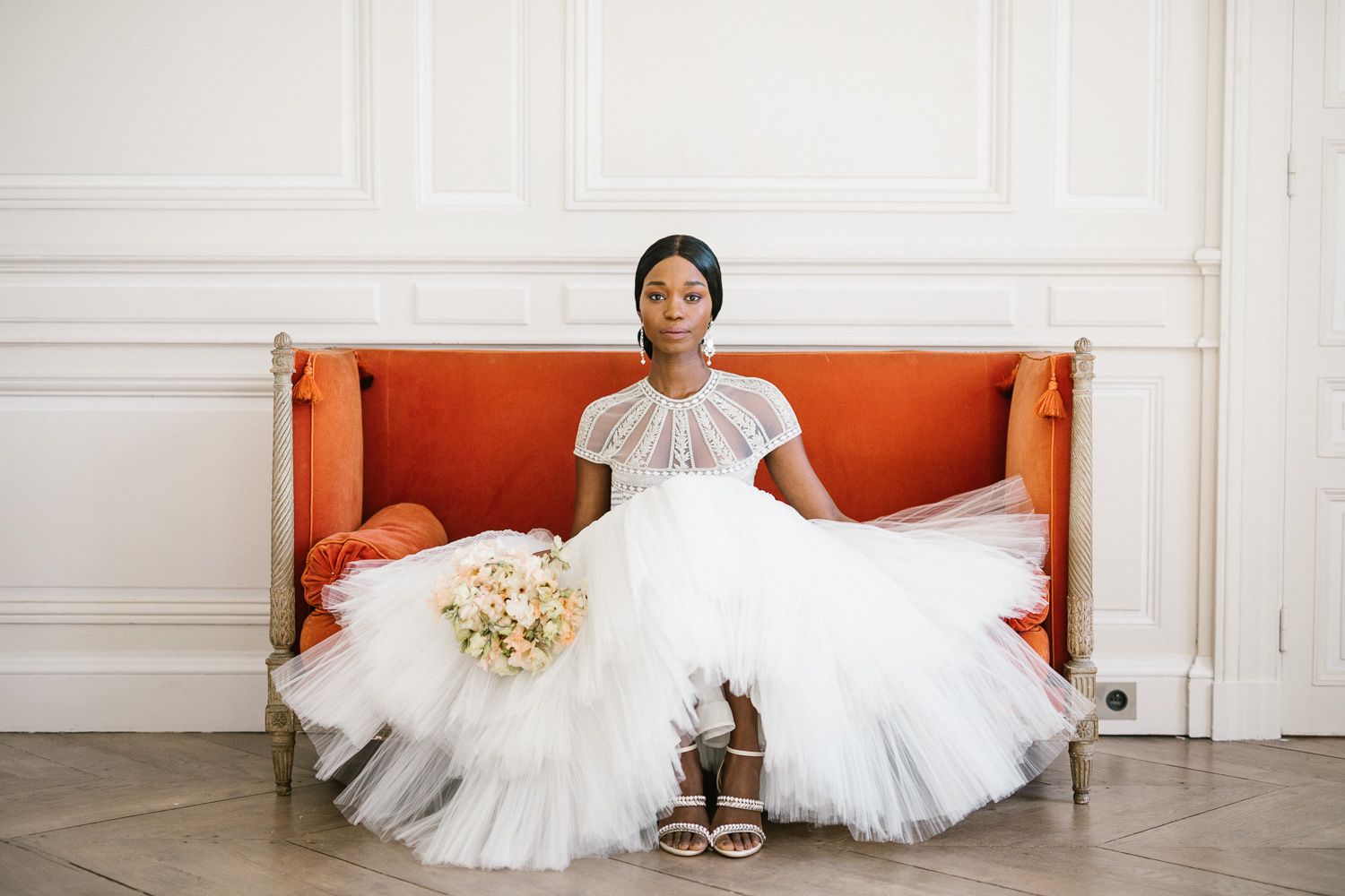 Bride in elegant white tulle gown sits on orange couch holding pastel bouquet in bright interior.