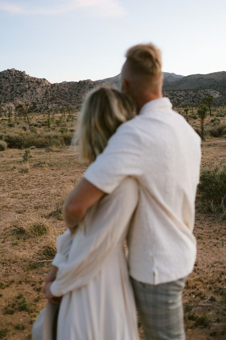 A couple embraces while looking out at a desert landscape with mountains and Joshua trees in the background.