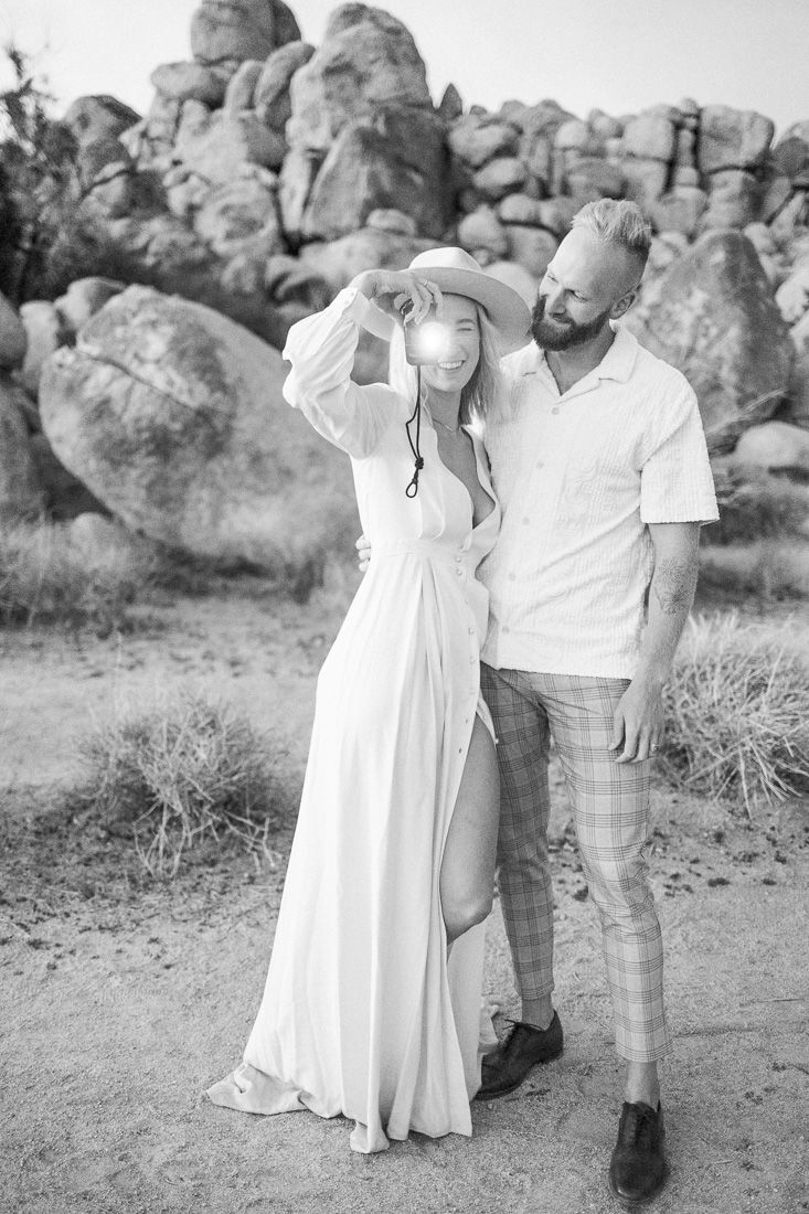 A stylish couple embraces near large desert boulders in coordinated white outfits.