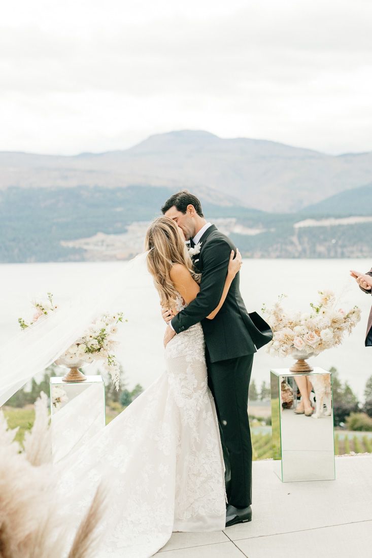 Newlyweds share an intimate kiss on a terrace overlooking a scenic mountain lake.