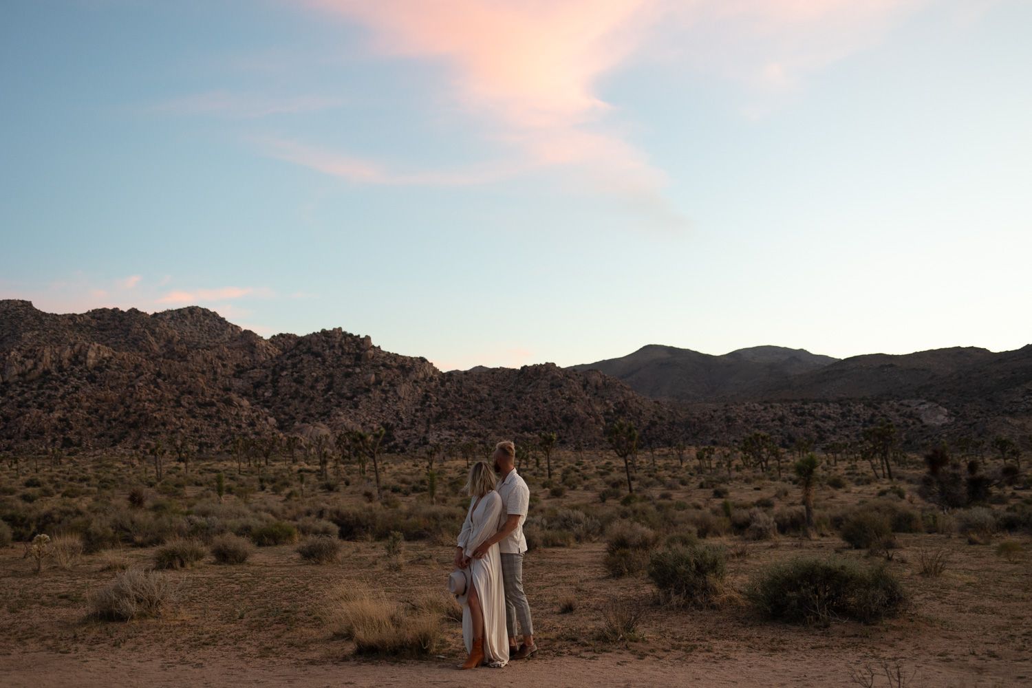A couple stands together against a desert mountain backdrop at sunset.