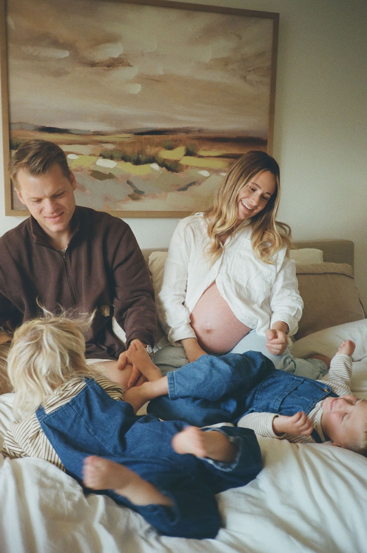 A family relaxes together on a bed, with a landscape painting hanging on the wall behind them.