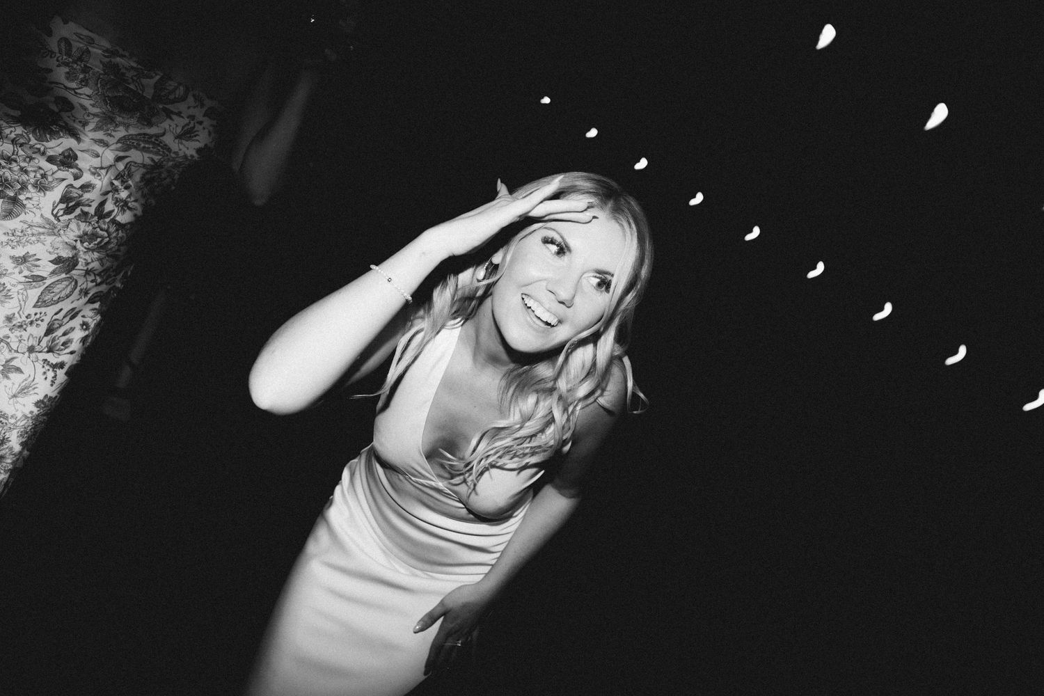 A person in a white dress smiles and dances at a party with confetti falling in black and white photography.
