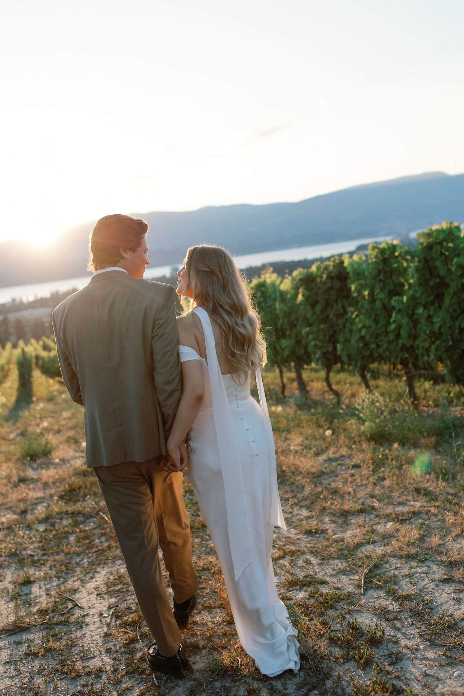 Couple in wedding attire standing together overlooking vineyard at sunset in mountainous landscape.