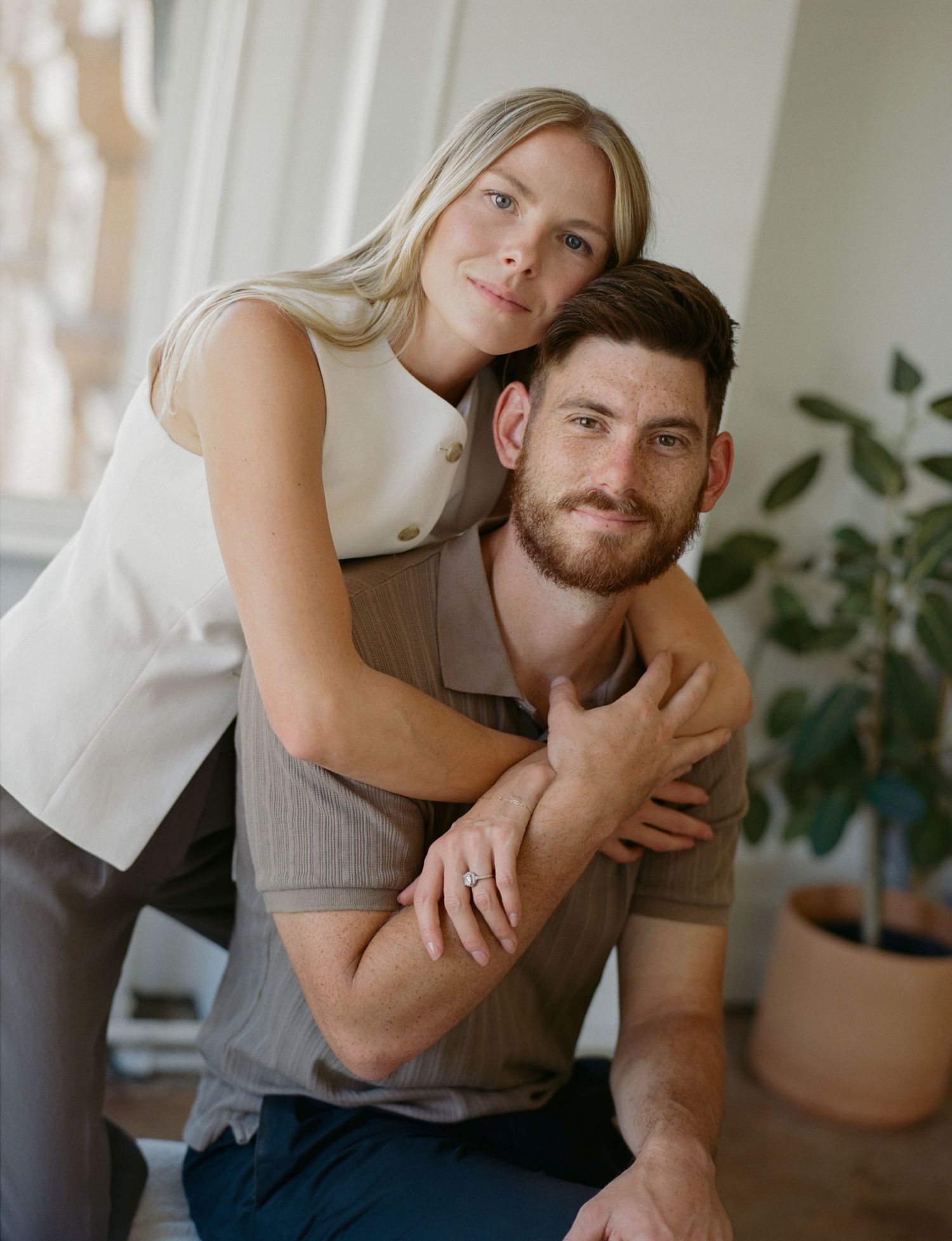 A couple poses intimately in a minimalist interior setting with houseplants and natural lighting.