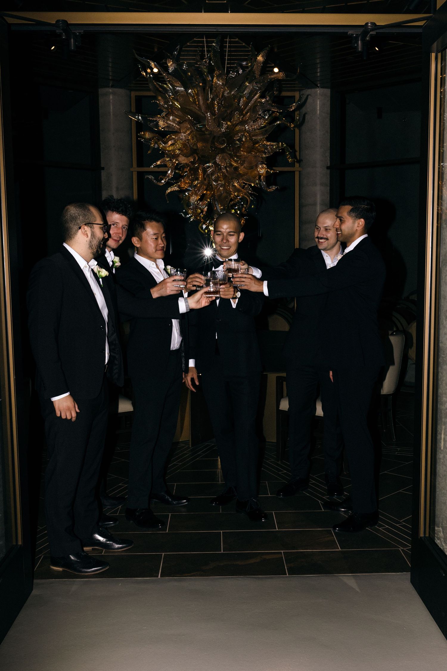 Group of men in black tuxedos socializing and toasting at a dimly lit event.