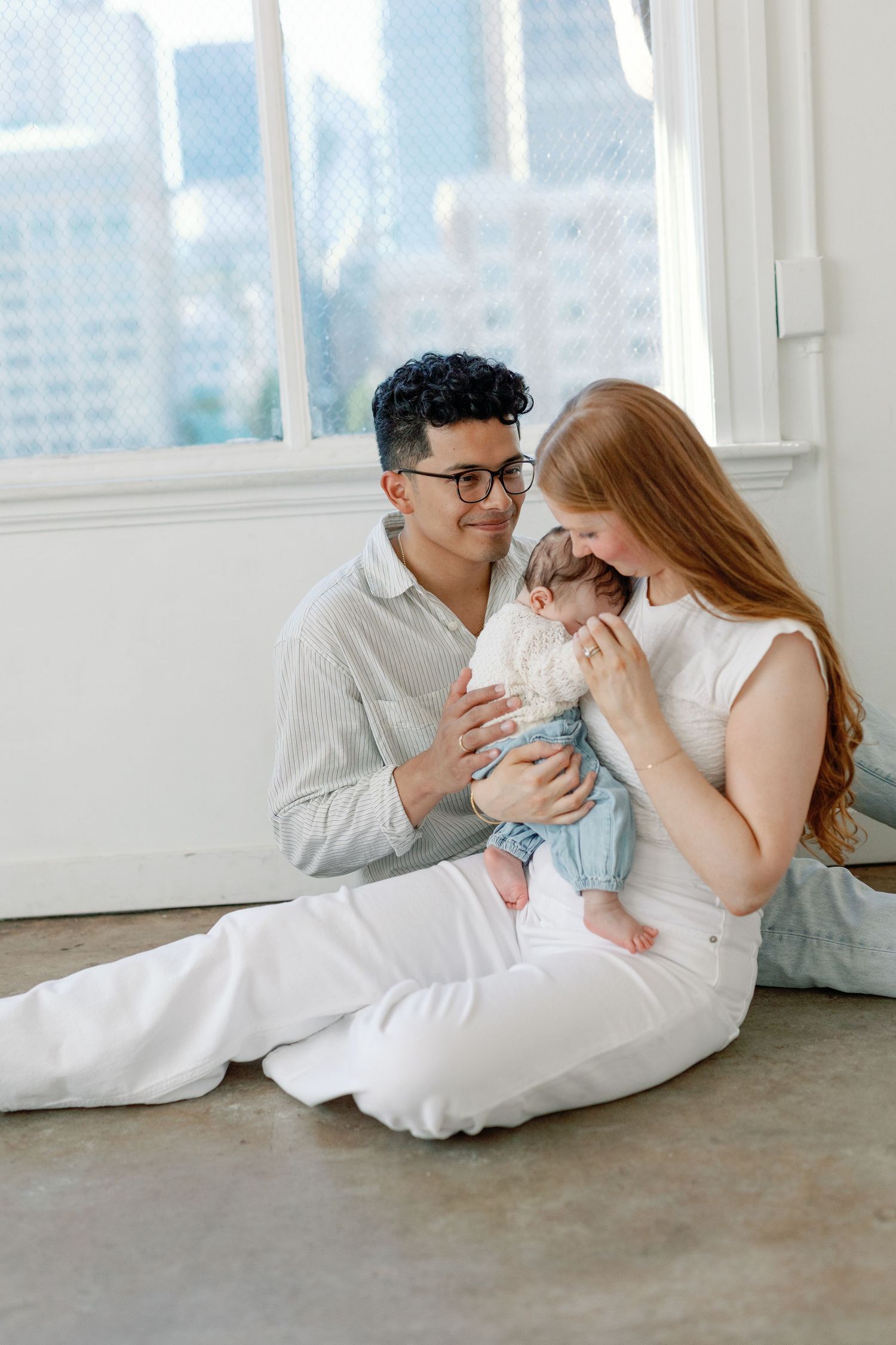 A couple in white clothing lies on the floor laughing and embracing in a bright room with city views.