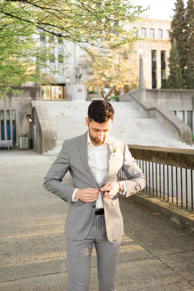 A groom adjusts his suit jacket buttons while standing on an outdoor walkway.