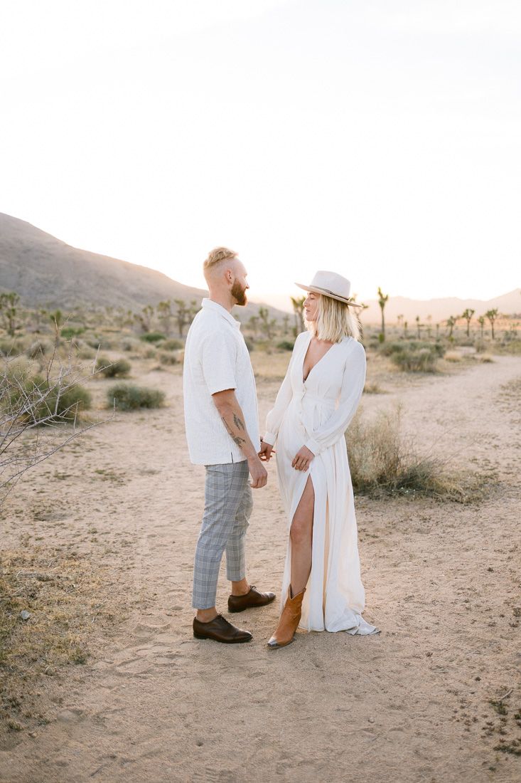 A couple shares an intimate moment on a desert trail at sunset with mountains and Joshua trees in the background.