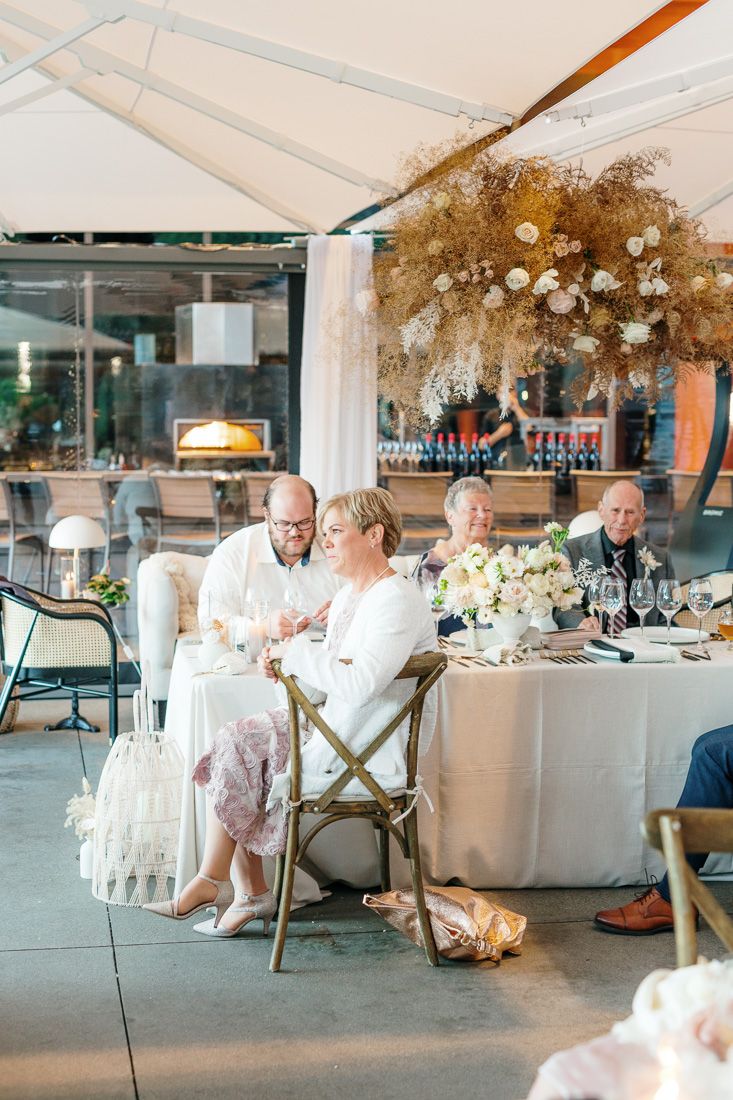 Wedding reception guests seated at tables under a white tent with elegant floral arrangements.