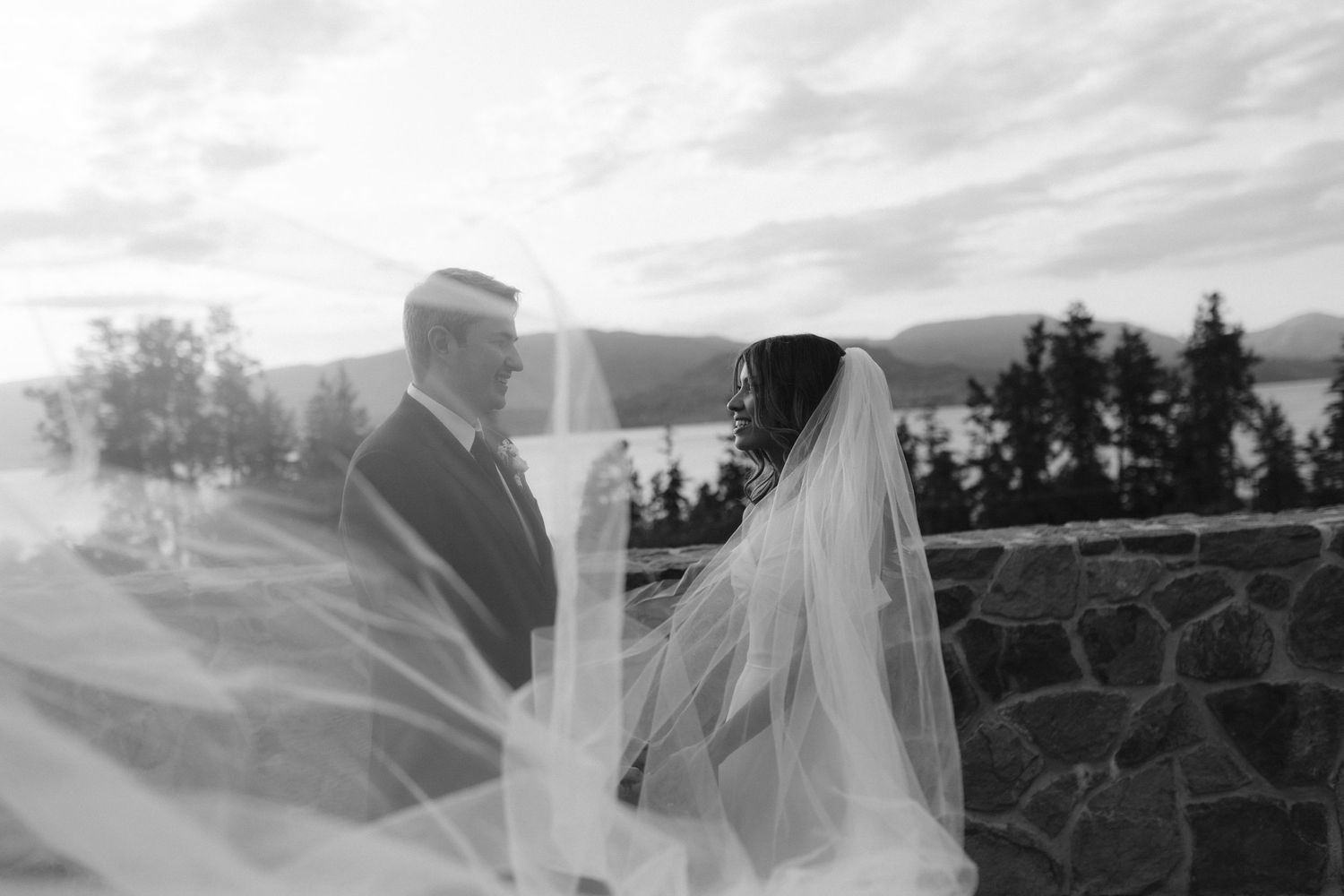 A black and white wedding photograph with a couple standing by a stone wall overlooking a scenic lake view.