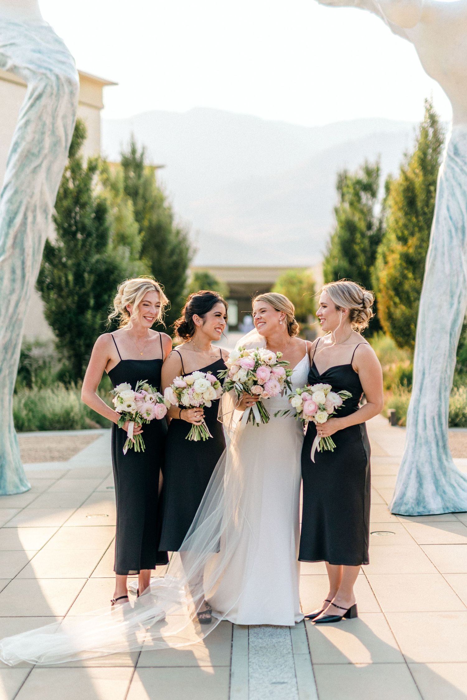 Wedding party poses outdoors with bridesmaids in black dresses holding pink bouquets beside a bride in white.