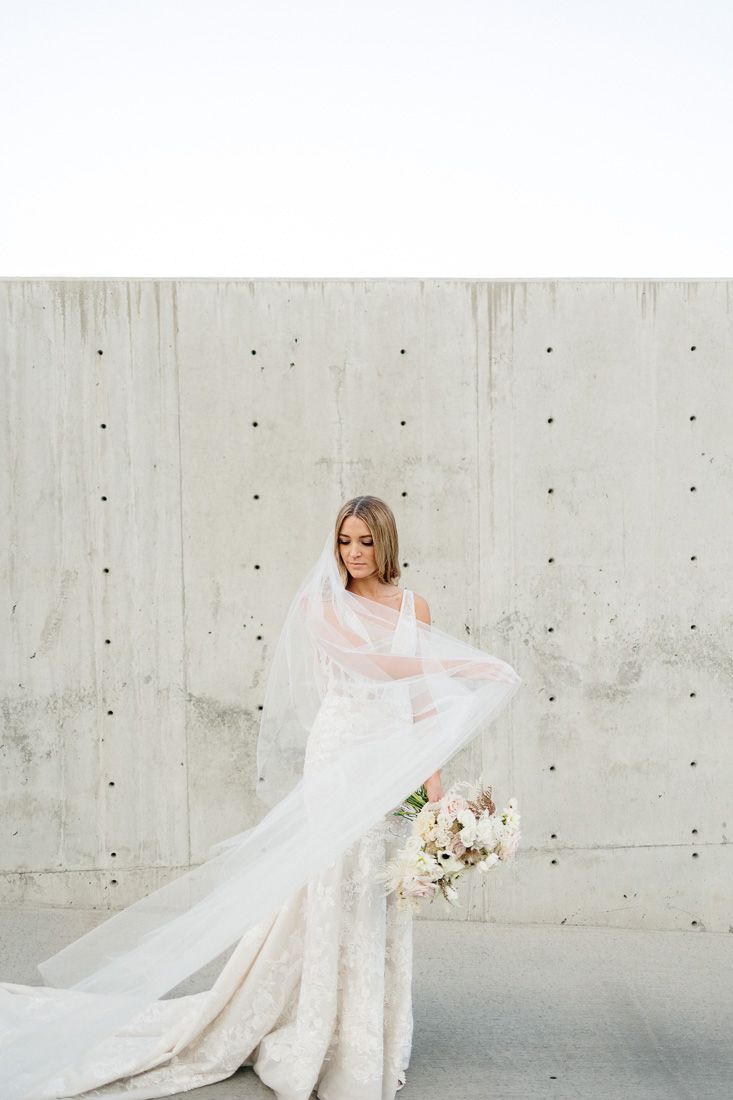 Bride in fitted lace gown with flowing veil poses against concrete wall.
