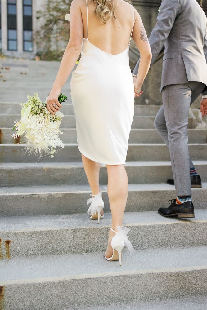 Elegant white cocktail dress and bow-adorned heels on stone steps with floral bouquet.