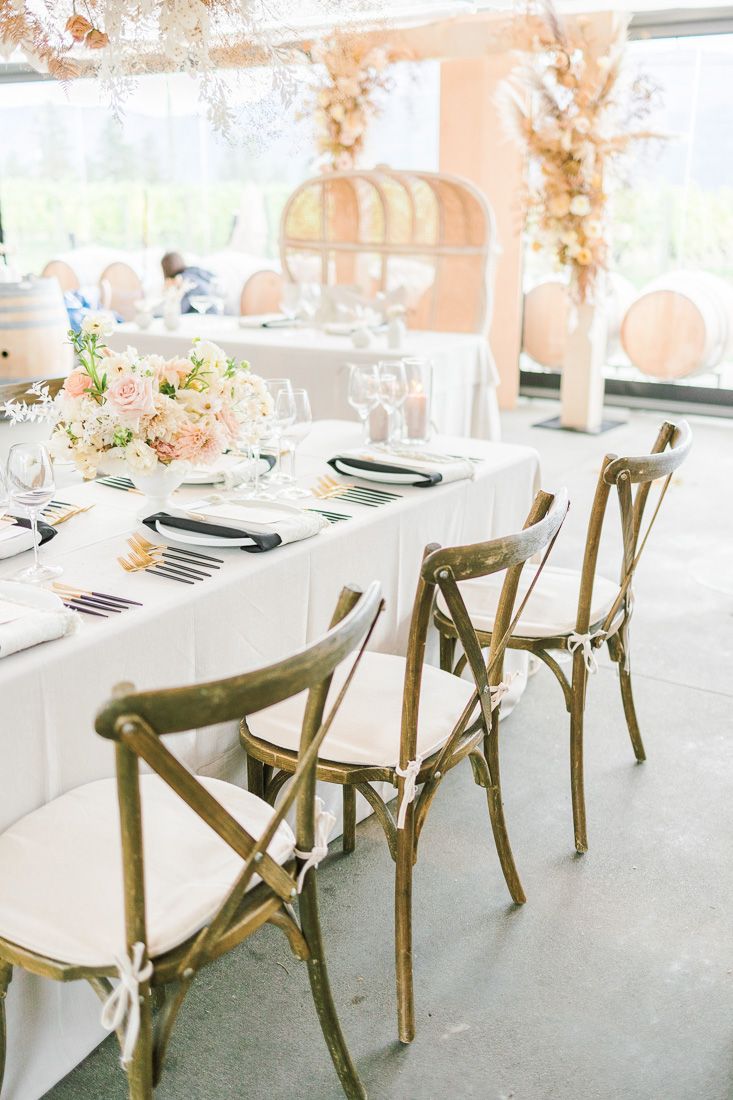 Elegant wedding reception table with white linens and rustic wooden cross-back chairs in a bright venue.