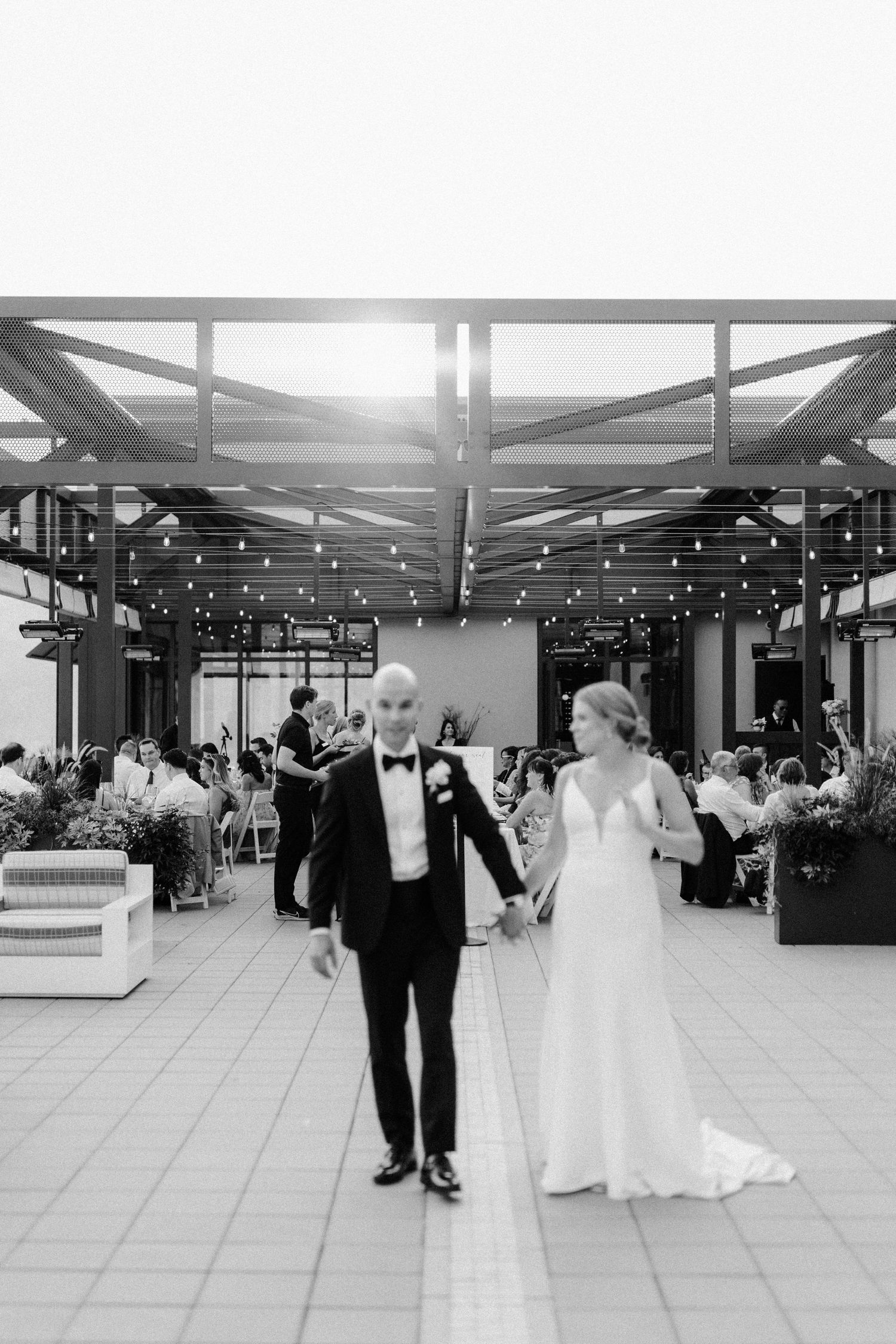 Black and white photo of newlyweds making their reception entrance under string lights and modern architecture.